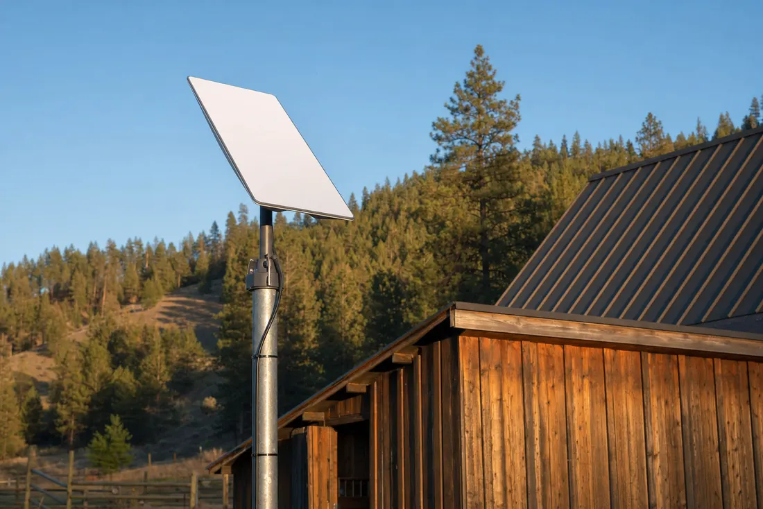 A satellite internet dish pole-mounted beside a rural barn with forested hills behind